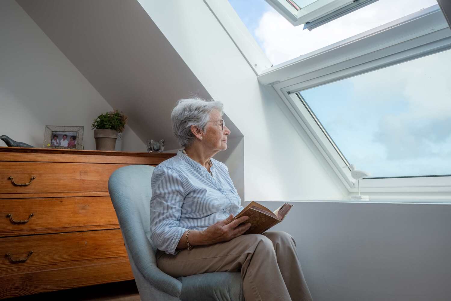 personne âgée avec un livre dans les mains, assise dans un fauteuil qui regarde le ciel bleu à travers sa fenêtre de toit ouverte